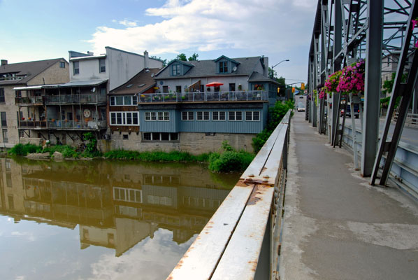 buildings along the river bank are reflected in the water seen from the bridge in Elora